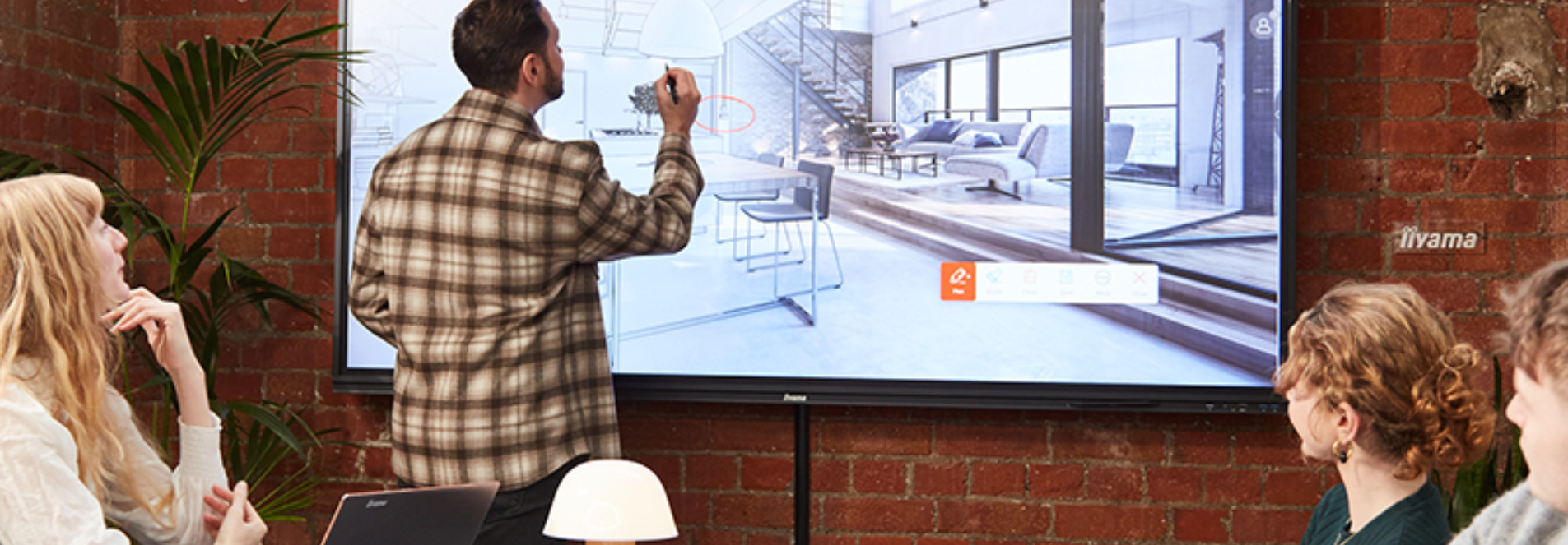A man stands before an iiyama interactive screen and writes on it while his colleagues sit at a table and watch. 