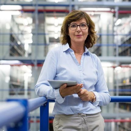 Woman smiles facing the camera, she stands in a factory setting holding a tablet.