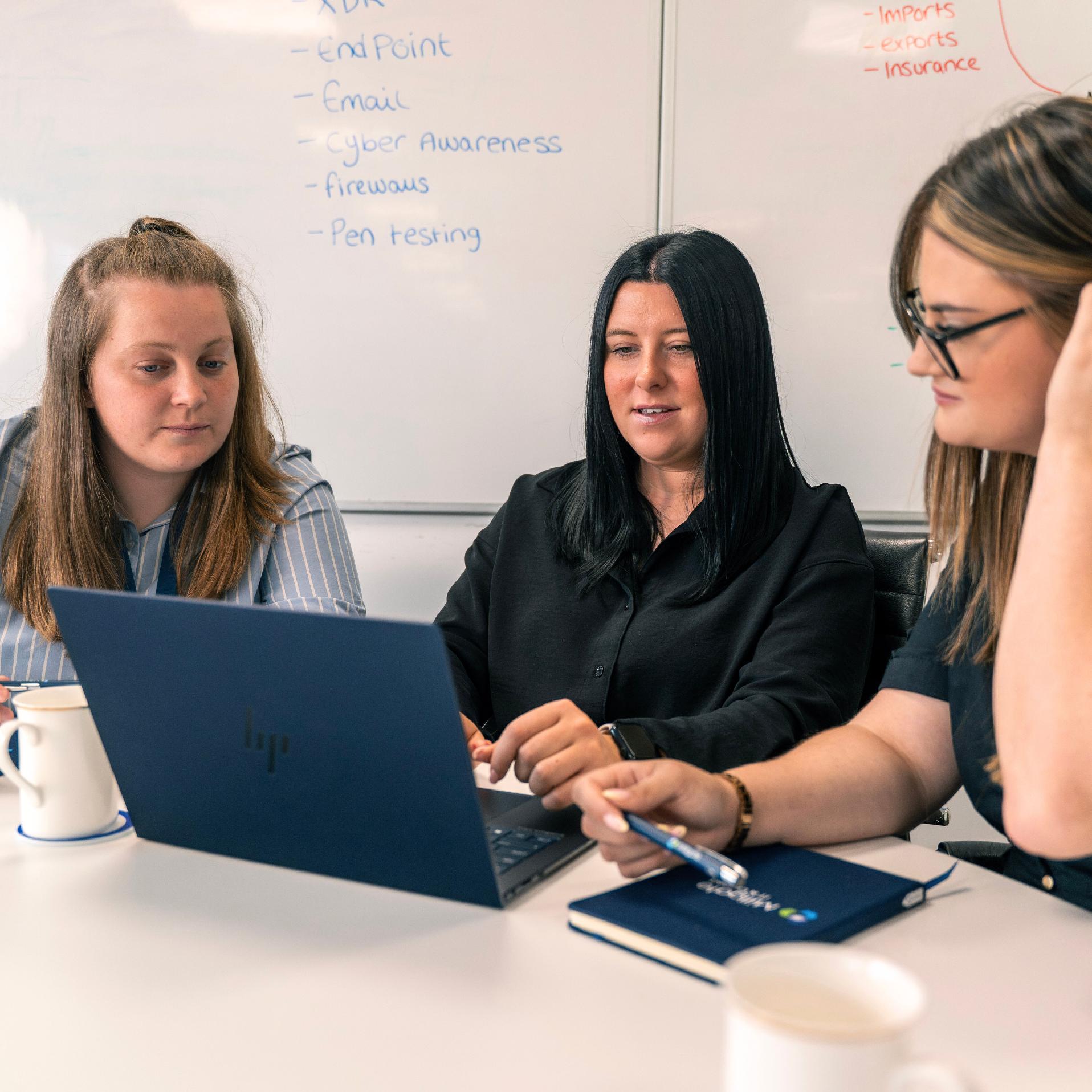 Three Millgate staff members work together in a meeting room around a single laptop. They appear focused on the t