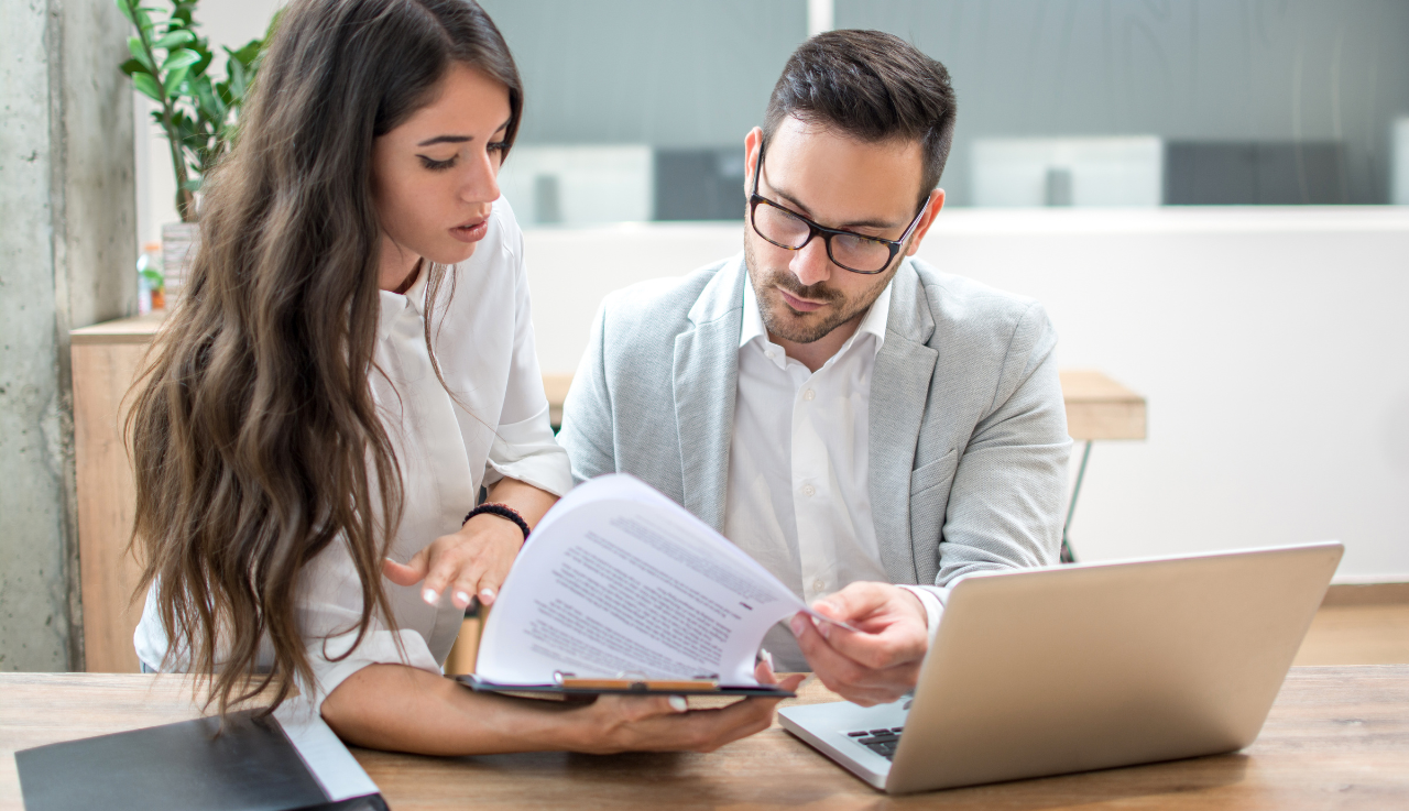 Two employees work together, one is holding a clipboard, the other is looking over it with a laptop in front of them.