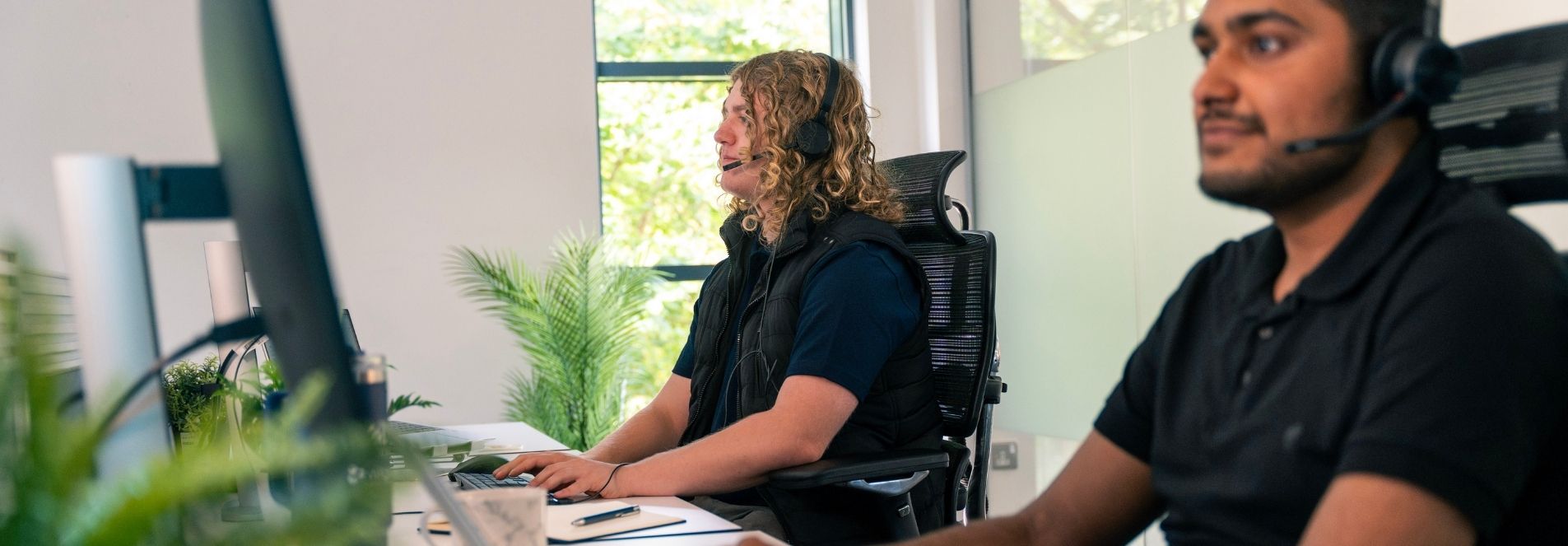 Two team members sit facing their monitors with headsets on.