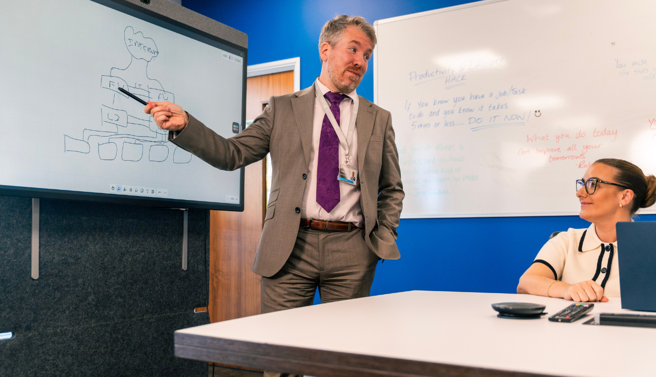 A staff member is presenting to another in a meeting room. He stands at the head of the table gesturing to the board behind him.