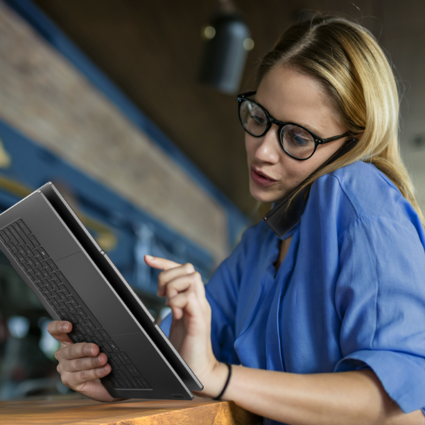 A woman is using a foldable laptop as she holds a phone between her shoulder and ear, she's on a call.
