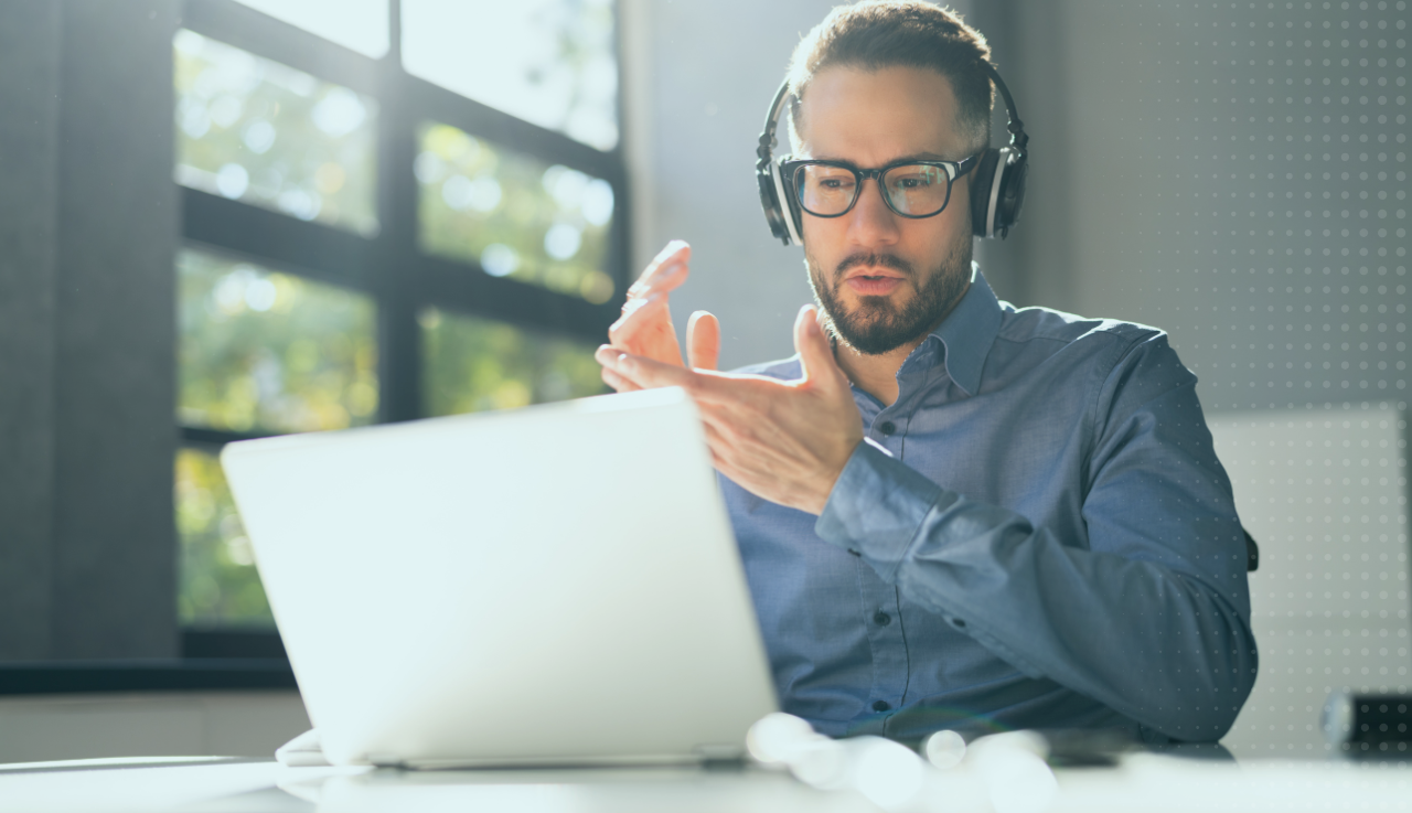 A man sits at his desk as he talks to someone on his laptop with a headset on.