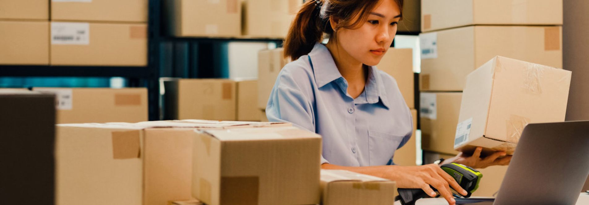 A woman is stood in a warehouse surrounded by boxes, she is working on a laptop, holding a box and a scanner.