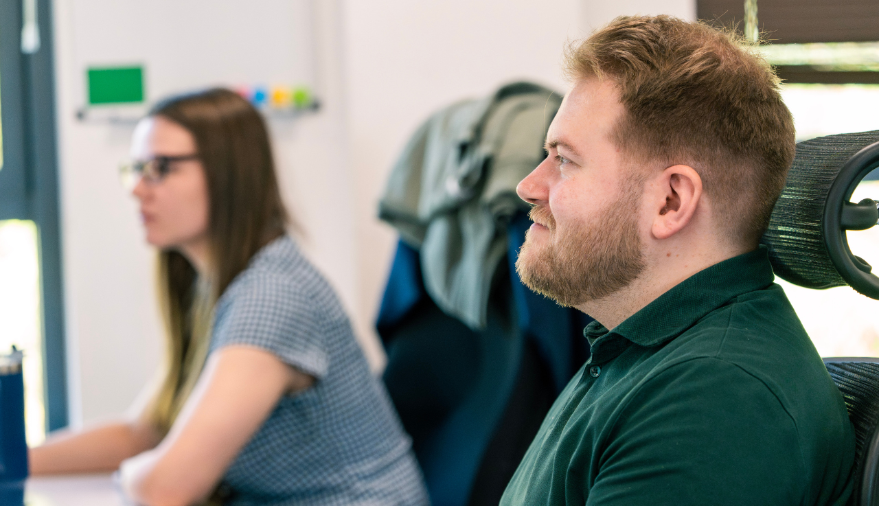 A team member at his desk. Another the same in the background, they are working.