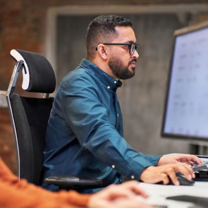 Man sat at desk with his hand on a mouse while he works on his desktop.