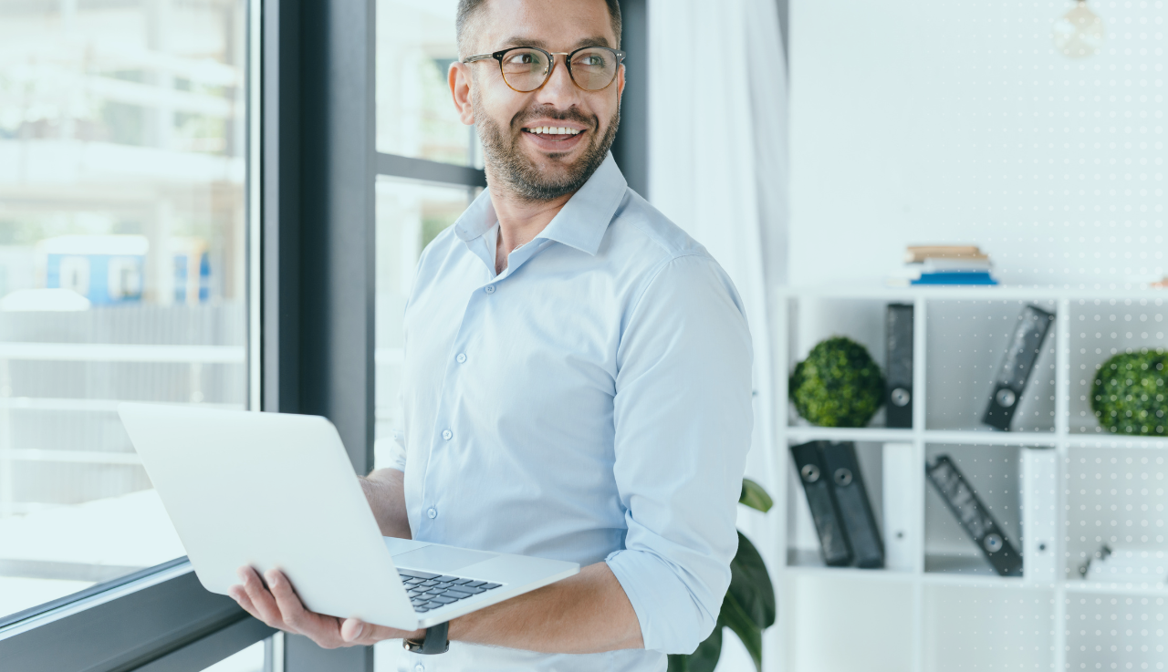 A man looks to the right smiling while he hold a laptop. He stand by a window.