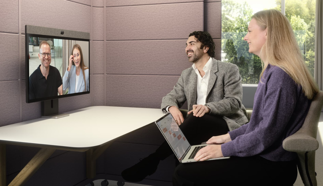 Two people sit at a table, one with a laptop on their lap. They look at a neat board where two more people are featured on the screen in a video call.