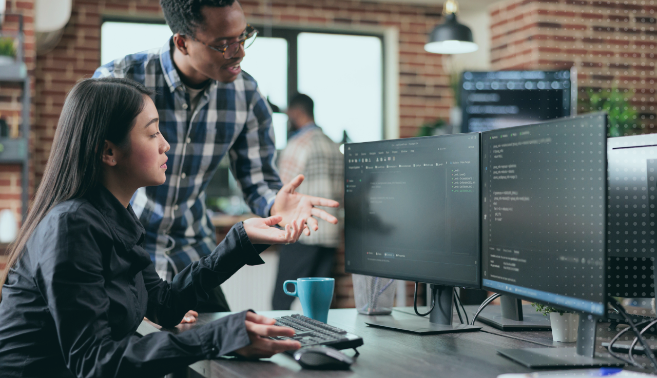 Woman sits at desk gesturing to her desktop screen, her colleague stands beside looking at the screen also.