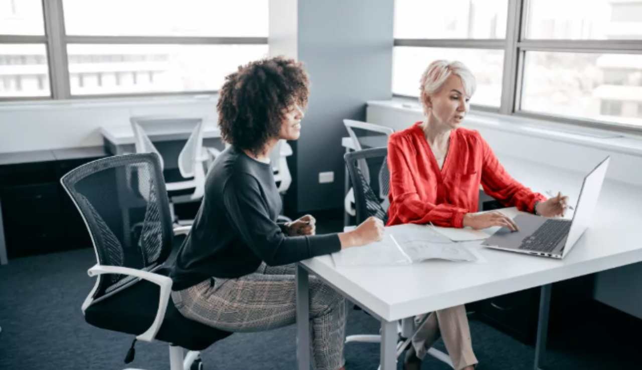 Two staff, female, sit at a desk together working from one laptop in an office space.