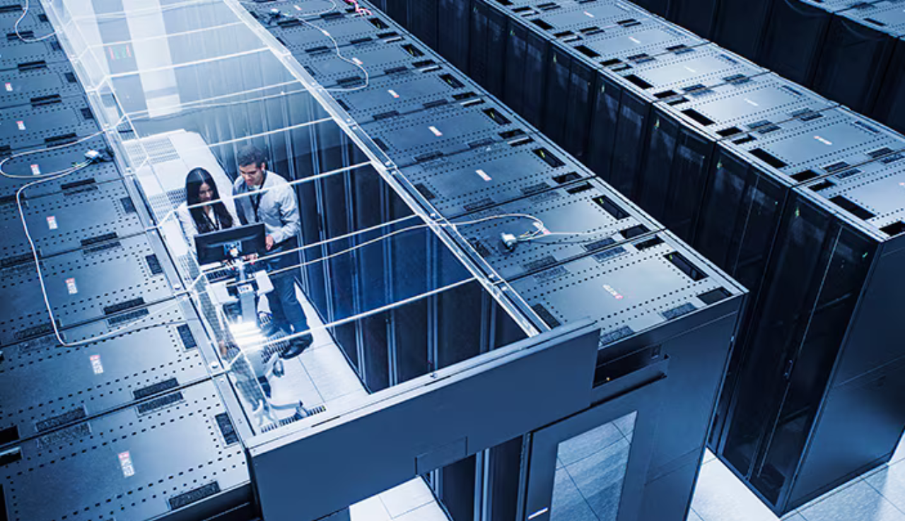 Overtop view of a large server room, two staff stand in one of the aisles looking at a laptop.