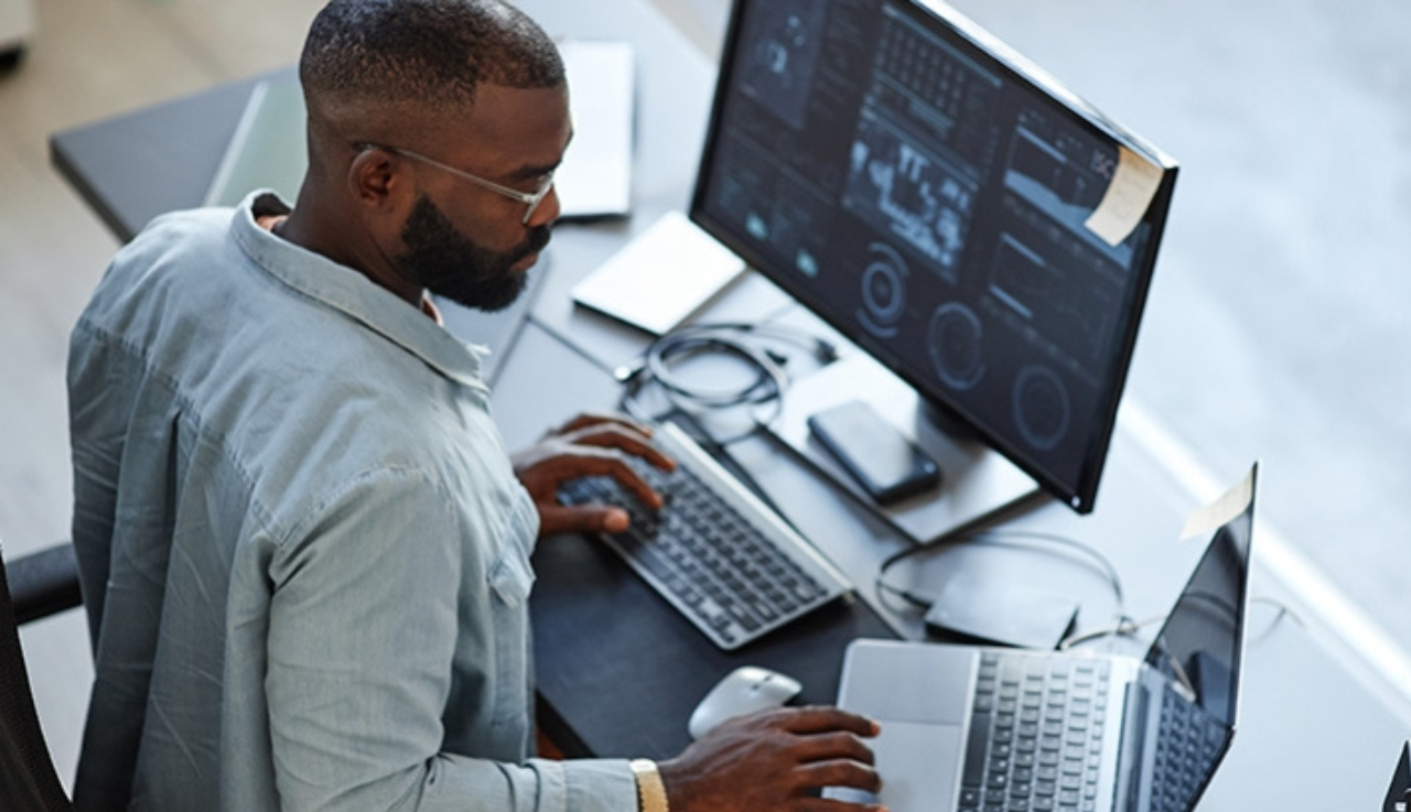 Man works at a desk with a laptop and a desktop.