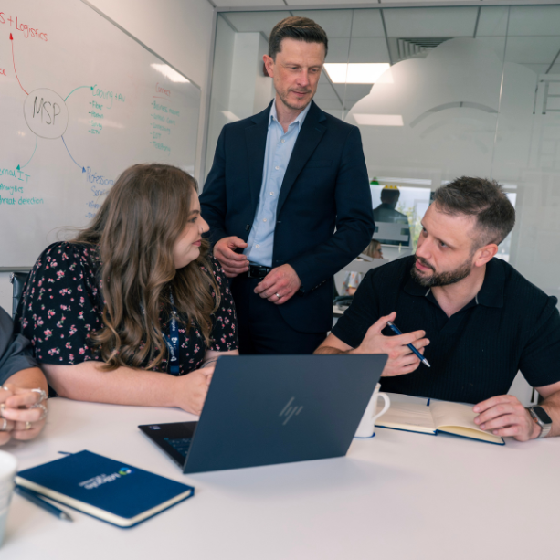 A group of three. Two sat, one standing. The work together at a table in the office, using a laptop and taking notes in a notebook.