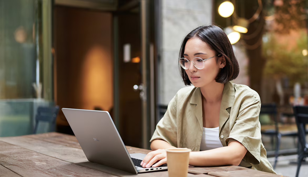 A woman sits a table outside wearing glasses. She is working on a Dell laptop.