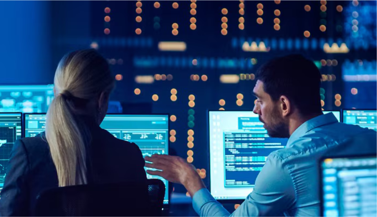 Two workers sit facing away at a couple of monitors. The room is blue and filled with lots of technical apparatus and blinking lights.
