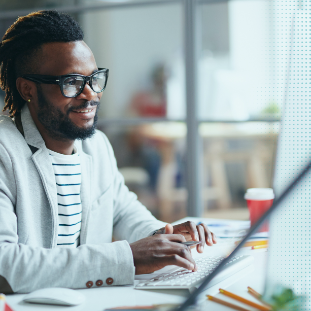 A man is facing right towards his desktop. He's typing on a keyboard and smiling.