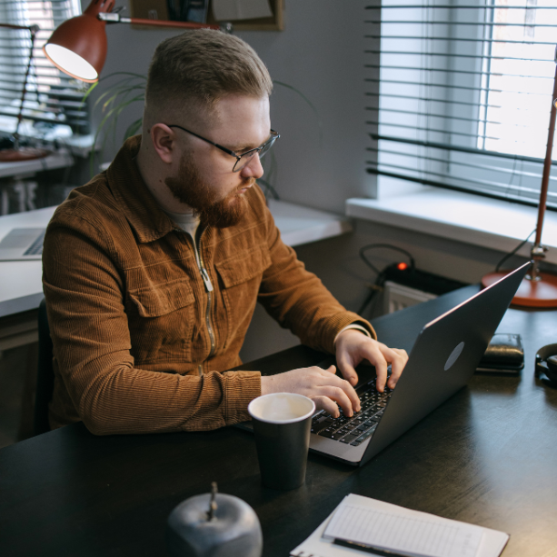 Man sits facing his laptop at a desk, he has a coffee cup beside him.