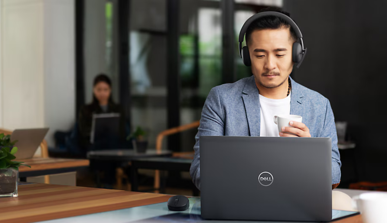 A man is sitting in an office holding a coffee while he works on a laptop at his desk.