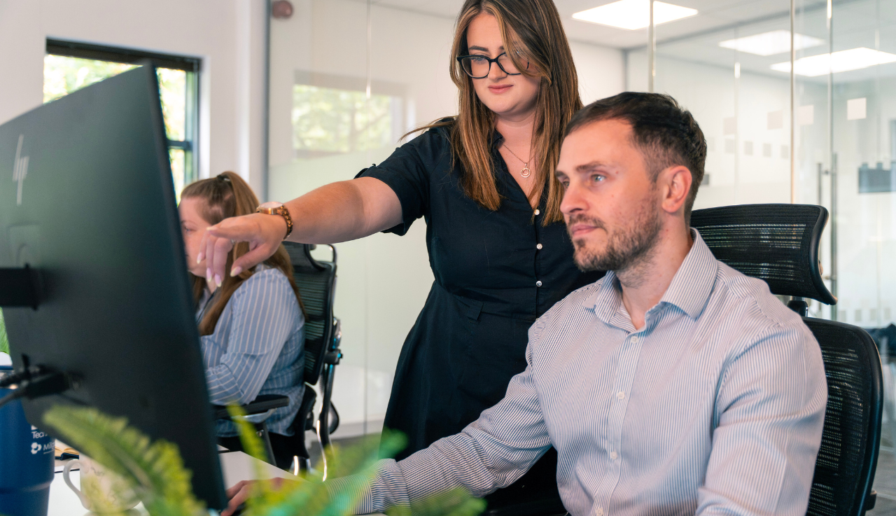 A female team member stands behind a male team member at his desk, she's pointing to his computer screen.