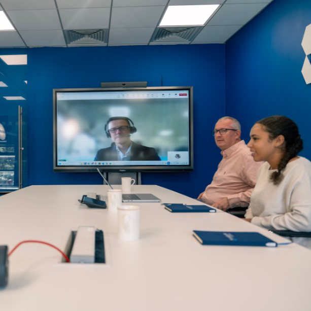 A meeting room with two staff at a table and another on a video call on the board.