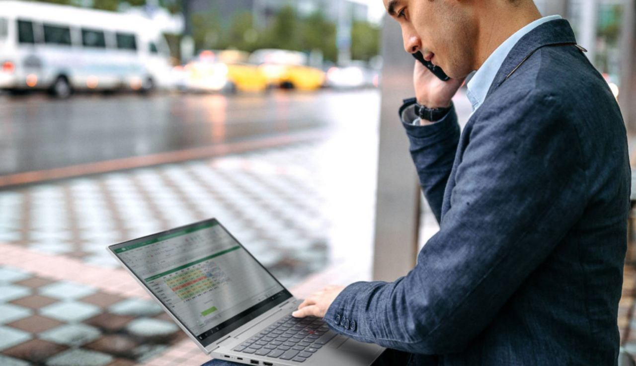 A man sits outside, a laptop on his lap as he talk on his phone whilst reading from his laptop screen.