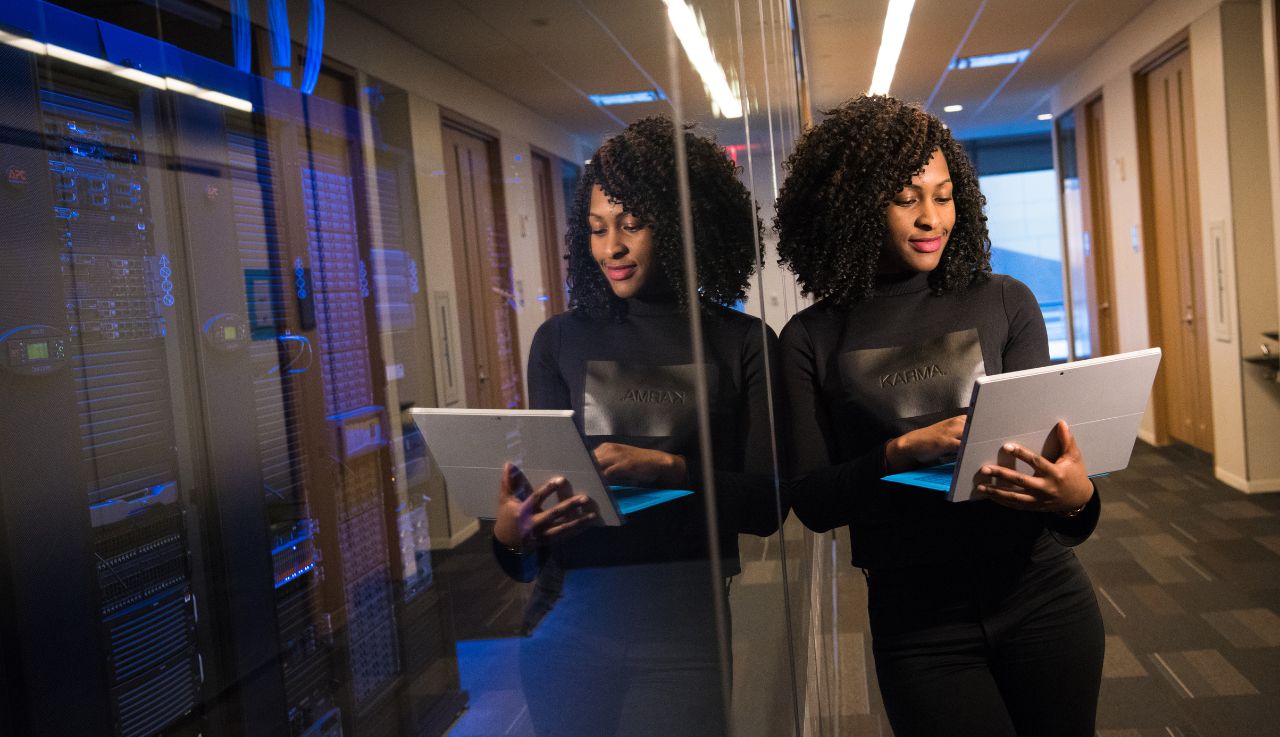 A woman stands by a glass wall that leads to a server room, she holds a laptop.