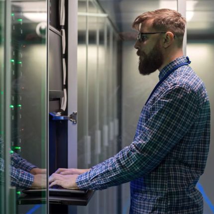 Man stands at a server cupboard as he types on the laptop within.