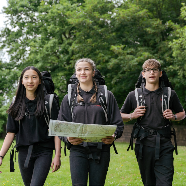 Three young people wearinf hiking gear walking through a field, the middle person holds a map.
