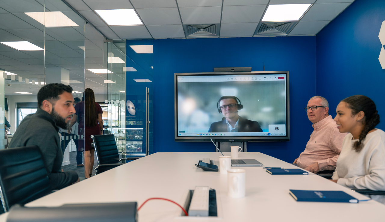 A meeting room with three team members seated at the table. In the background a smartboard shows another team member calling to the meeting.