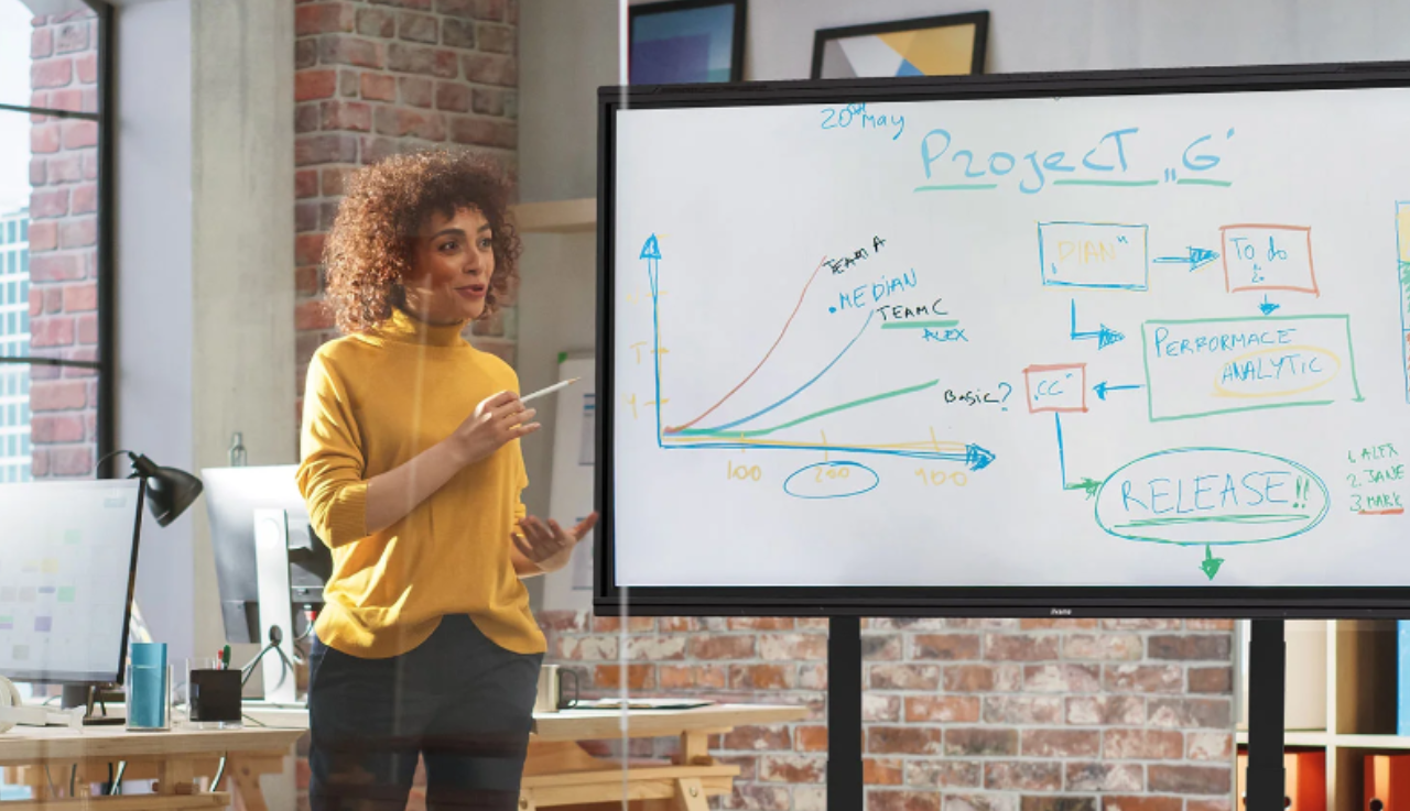 Woman stands in front of smartboard with info on it, she's presenting.