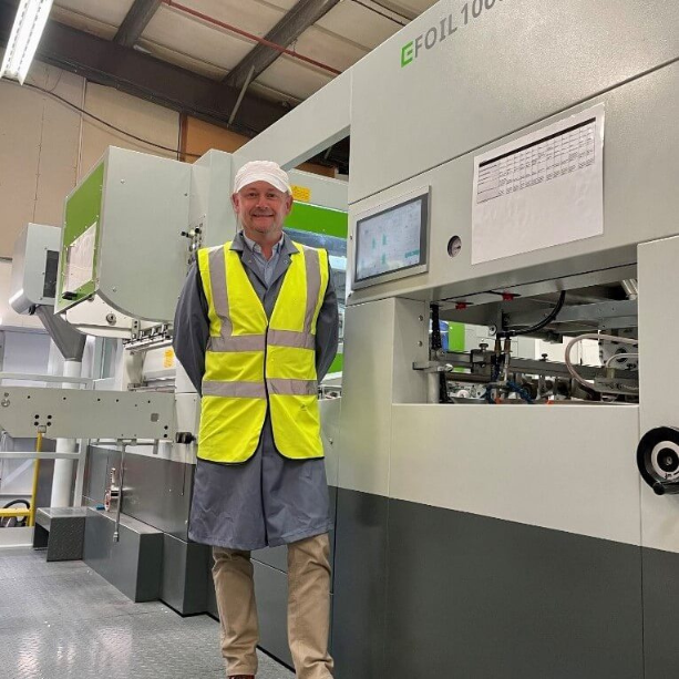 Staff in high-vis and hard hat in front of printing press equipment in factory.