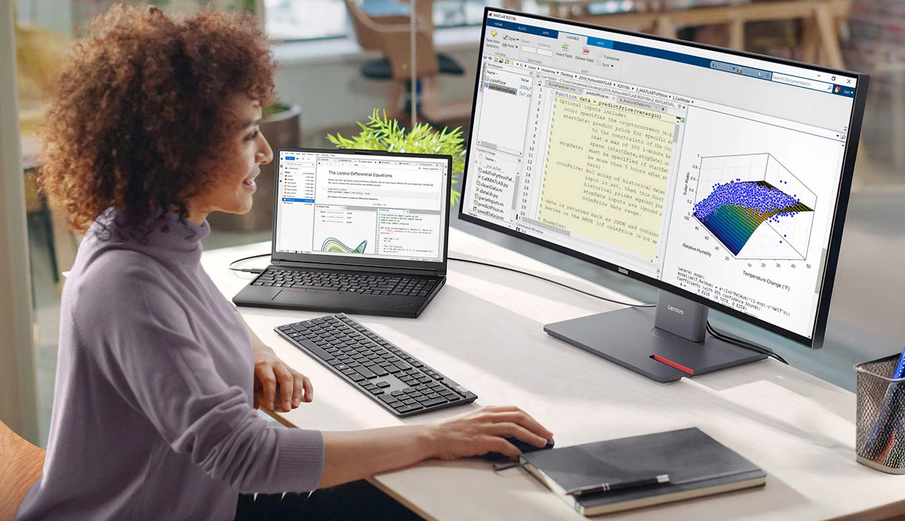 A woman is smiling facing her monitor and working on it as she sits at her desk. There's a laptop, keyboard and notebook on the desk.