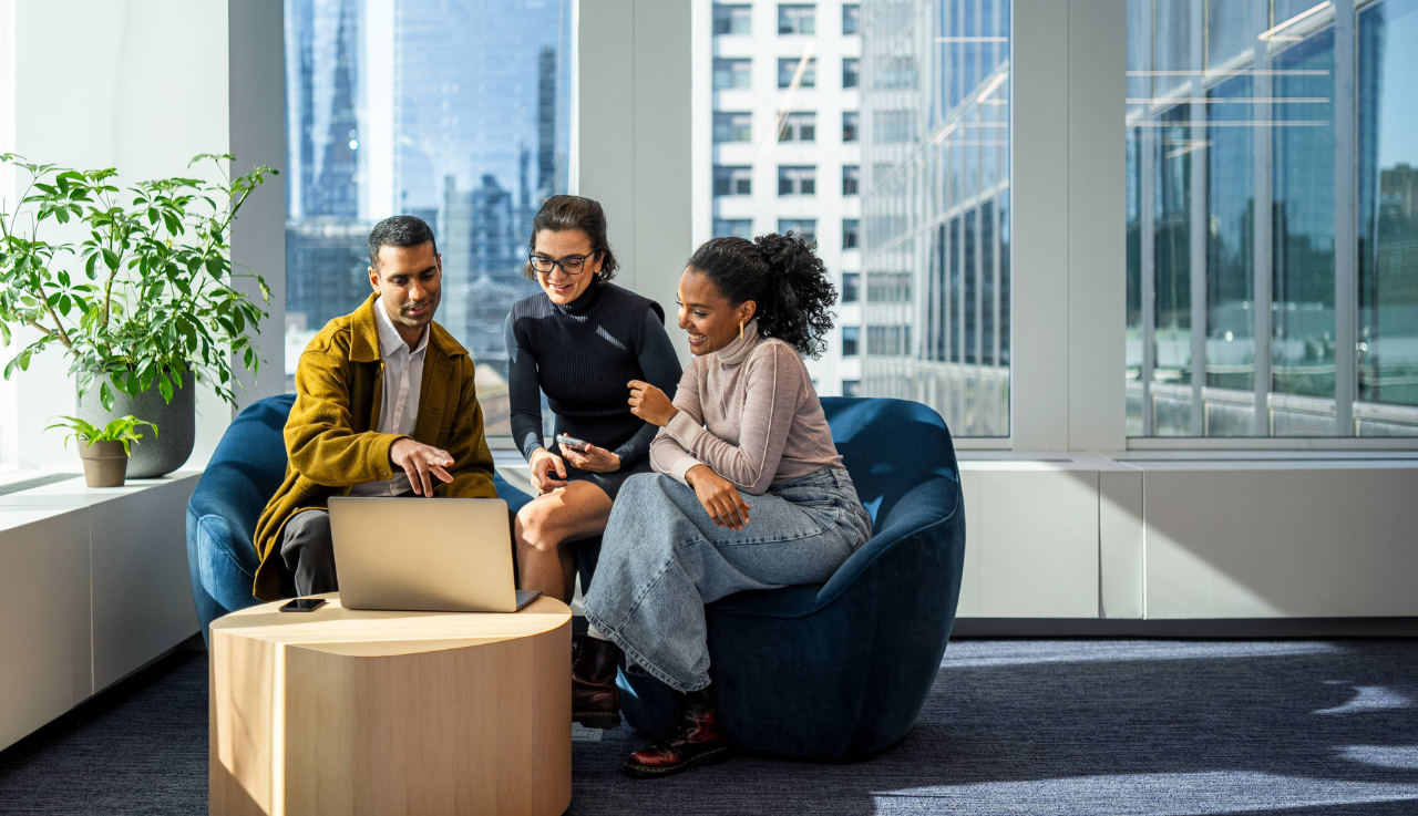 Three team members sat by a window on a sofa looking at a single laptop.