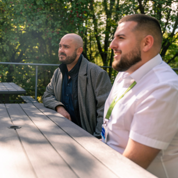 Two male team members sit outside together at a picnic bench talking.