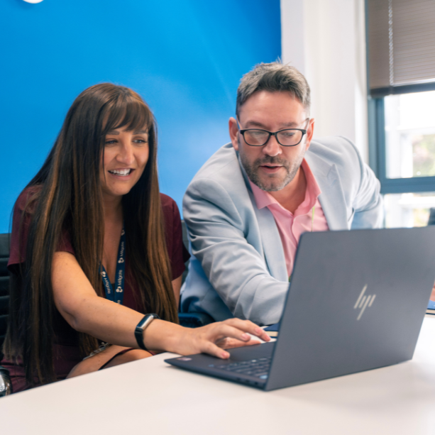 A male and female pair of team members sit together facing a HP laptop, smiling.