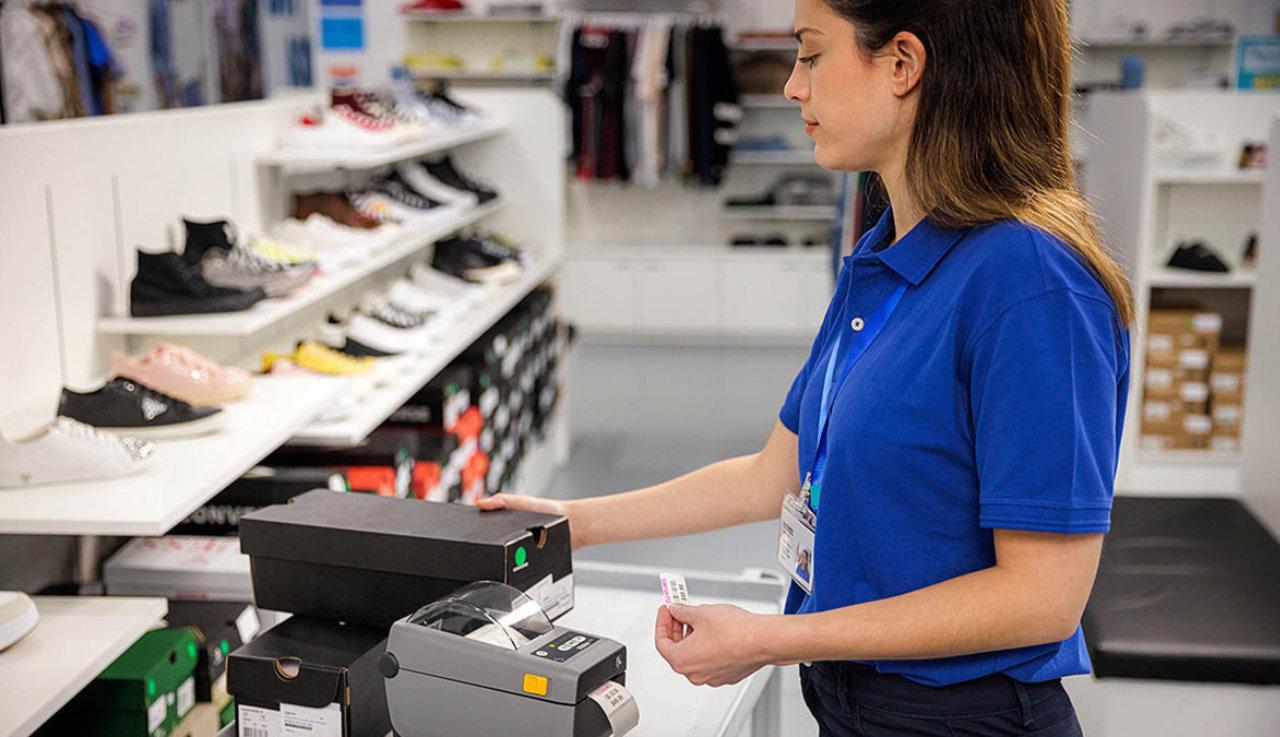 A shop worker is stood in a shoe shop holding a shoe box and a stands in front of a zebra label printer.